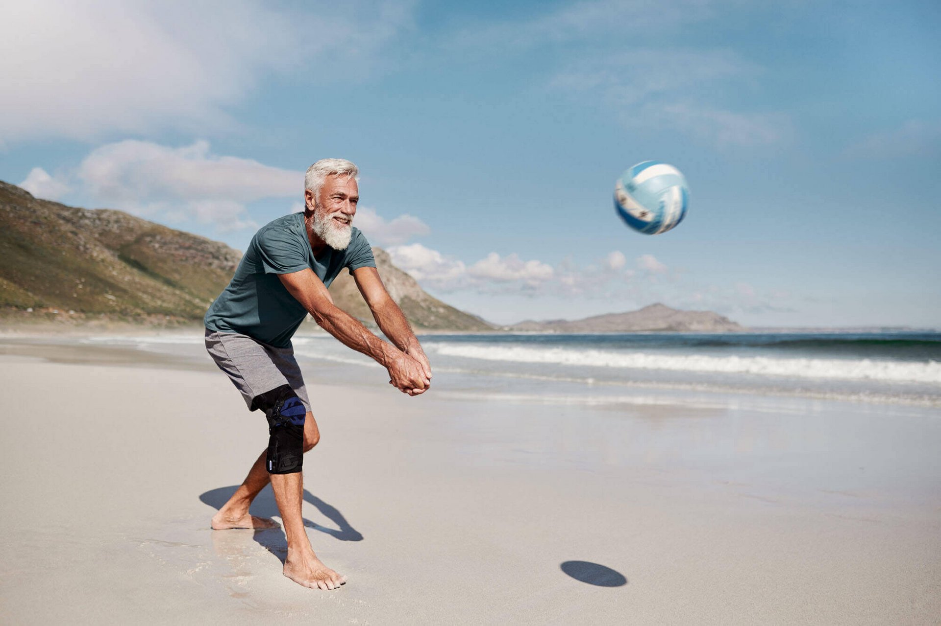 Ein Mann steht an einem Sandstrand und spielt einen Volleyball zurück. Er trägt eine GenuTrain OA Kniebandage von Bauerfeind für eine gezielte Entlastung und Stabilisierung bei Kniearthrose.